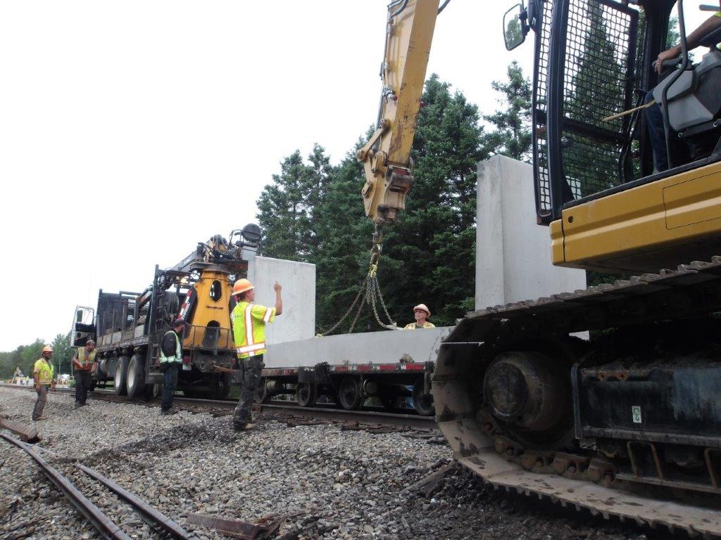 Workers in reflective vests and hard hats signal for the railroad ballast retainer to be lifted off of a train car